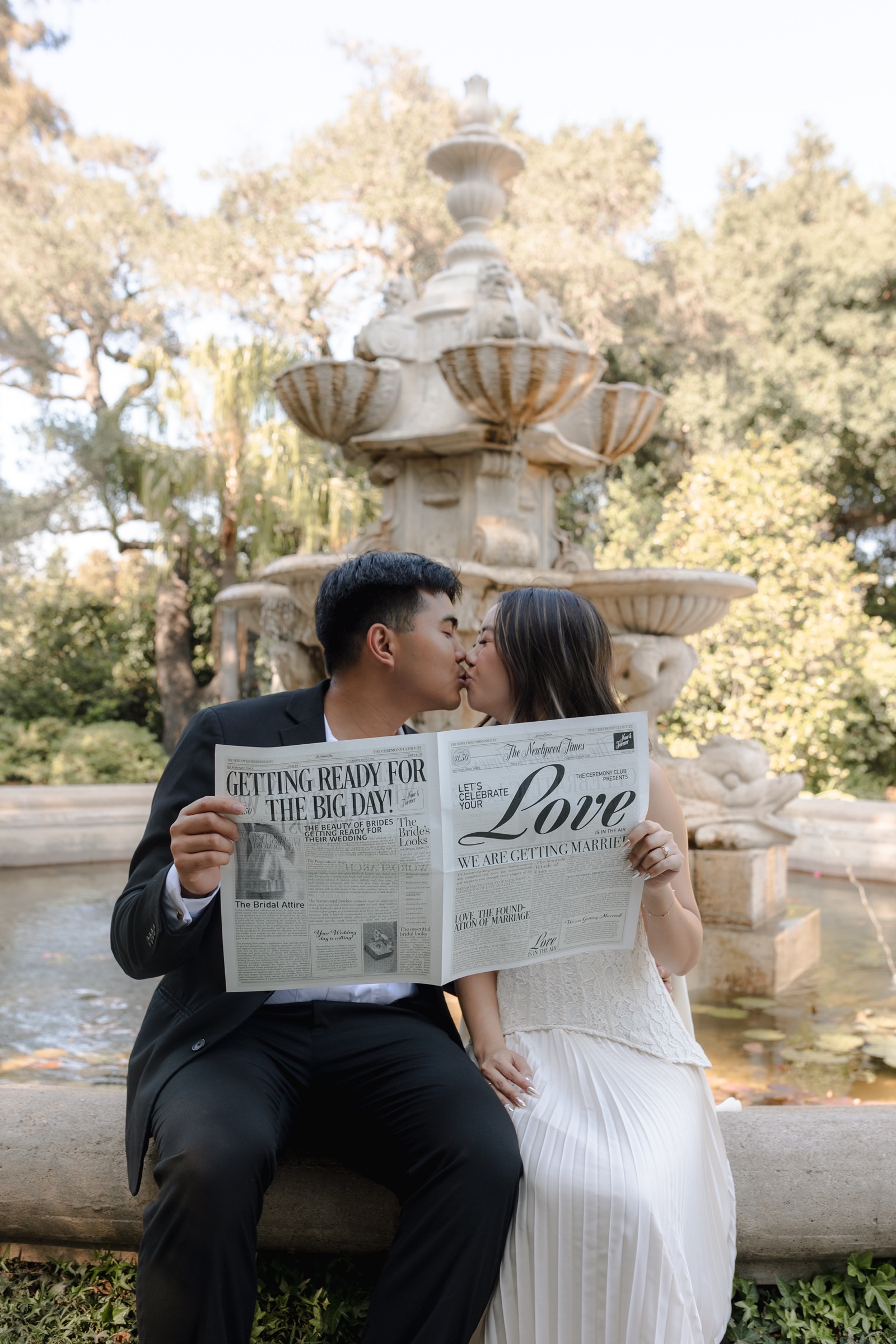 Edwin and Grace holding a wedding newspaper by the fountain during their engagement shoot